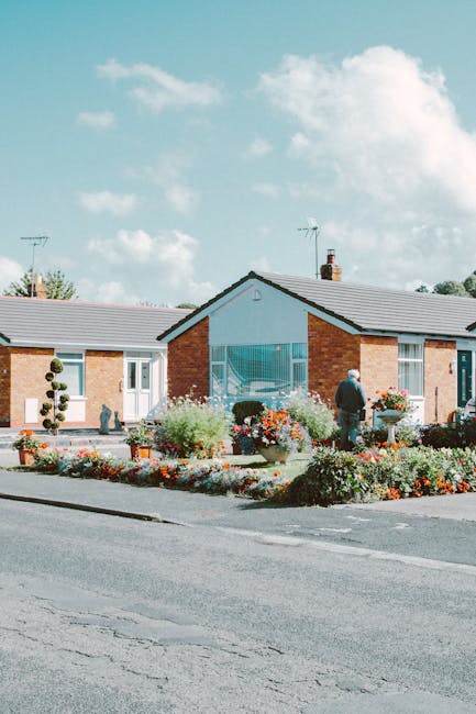 A neatly maintained front garden in a residential area featuring a low brick building with a large central window. The garden beds are filled with vibrant, blooming flowers in red, pink, and white, bo