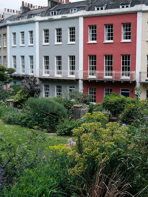 A neatly maintained front garden in a residential area featuring a low brick building with a large central window. The garden beds are filled with vibrant, blooming flowers in red, pink, and white, bo